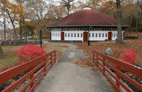 A round building with a red roof surrounded by autumn foliage and a small red bridge leading to it.