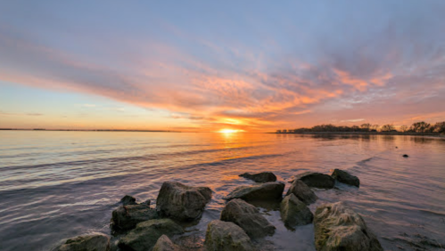 A serene sunset over calm waters, with colorful clouds and rocky shoreline in the foreground.