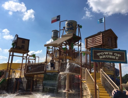 Water play structure with slides and water features, featuring a "Town of Brushy" sign and Texas flags under a blue sky.