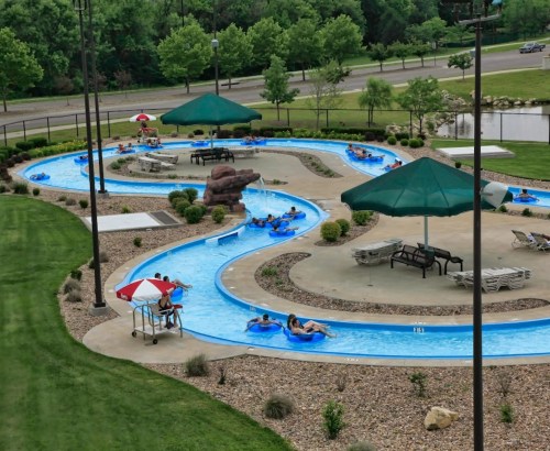 A winding lazy river pool with people floating on tubes, surrounded by green trees and shaded seating areas.