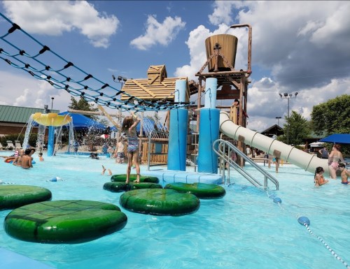A vibrant water park scene with kids playing on lily pads, a water slide, and a large play structure under a blue sky.
