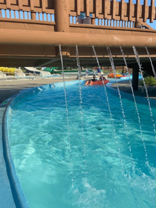 A clear blue pool with water cascading from a wooden structure above, surrounded by lounge chairs and greenery.