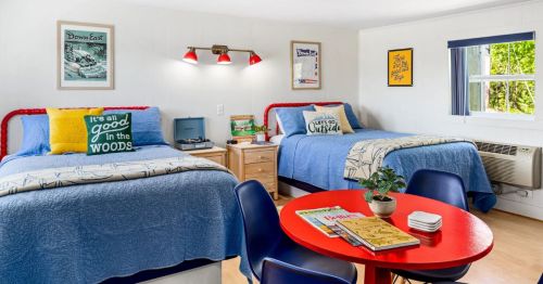 A bright and airy motel room with two queen beds made up with blue comforters and kitschy pillows