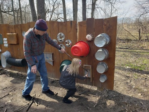 An adult and a child interact with a wooden wall featuring various metal objects and instruments in a natural setting.