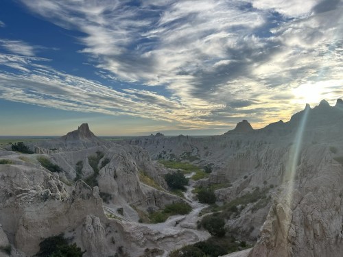 A scenic view of rugged, eroded rock formations under a cloudy sky at sunset, with green vegetation in the valley.