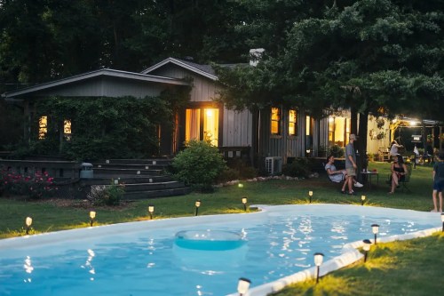 A serene evening scene with a glowing pool, cozy house, and people enjoying the outdoors in a lush garden.