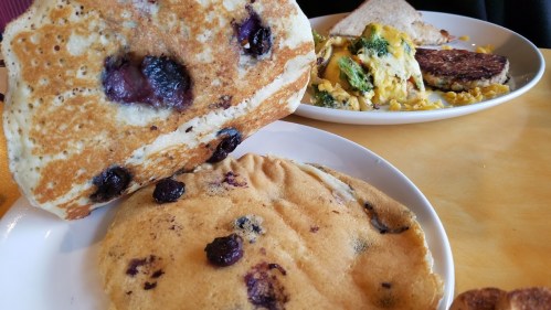 A stack of blueberry pancakes on a plate, with a side of scrambled eggs and a veggie patty in the background.