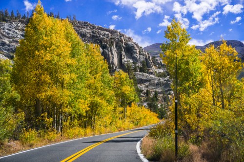 A winding road surrounded by vibrant autumn trees and rocky cliffs under a blue sky with fluffy clouds.