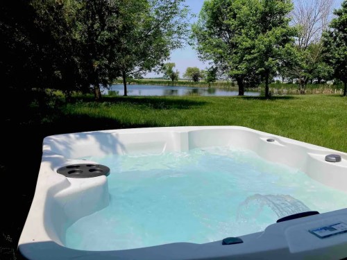 A hot tub with clear water sits in a grassy area near a serene lake surrounded by trees on a sunny day.