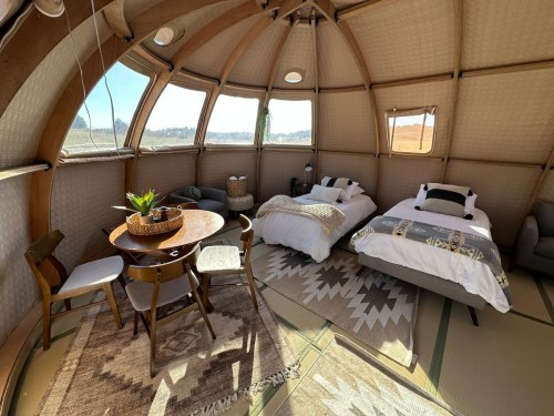 Cozy interior of a yurt featuring two beds, a dining table, and natural decor, surrounded by large windows.