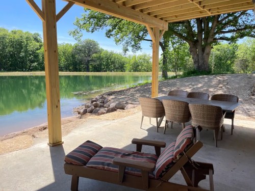 A shaded patio by a serene pond, featuring a table and chairs, with lush greenery in the background.