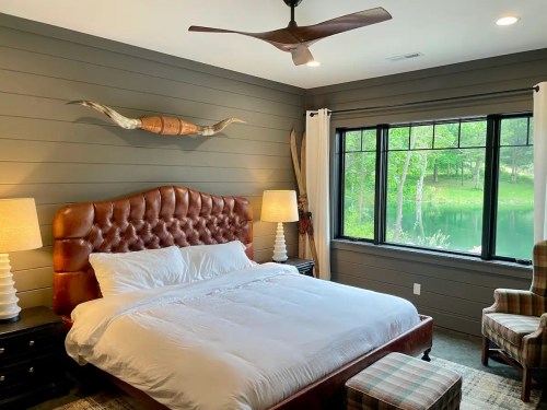 Cozy bedroom with a tufted leather bed, large windows overlooking greenery, and stylish decor.