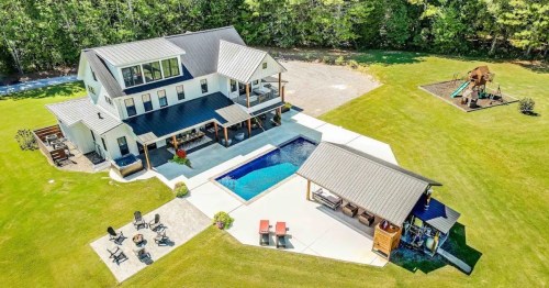 Aerial view of a modern home with a pool, outdoor seating, and a playground in a spacious green yard.
