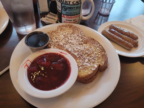 A plate of French toast dusted with powdered sugar, served with strawberry topping and a side of sausage links.