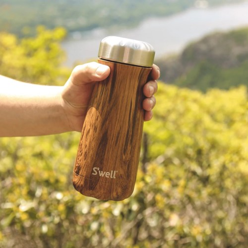 A person holds a S'well water bottle with a wood-like finish against a scenic outdoor backdrop.