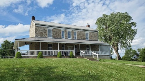 A two-story stone house with a porch, surrounded by green grass and trees under a partly cloudy sky.