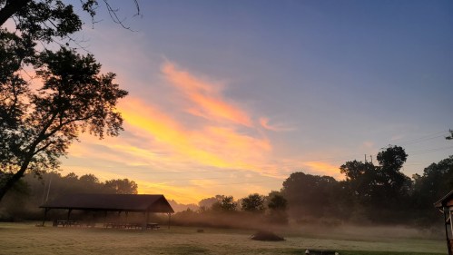 A serene sunrise over a misty field, with a pavilion and trees silhouetted against a colorful sky.