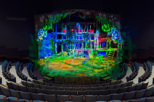 A vibrant theater stage set with colorful lighting and greenery, viewed from the audience seating area.