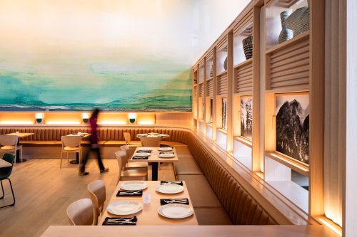 A modern restaurant interior with wooden seating, plates set on tables, and a large abstract mural on the wall.