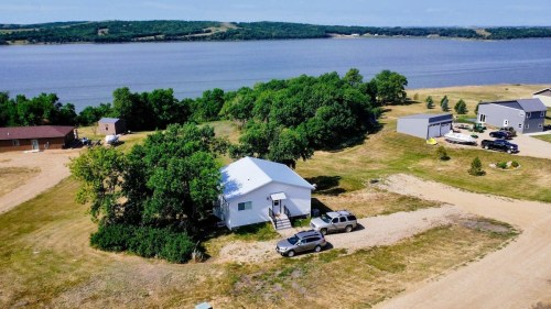 Aerial view of a house surrounded by trees, with vehicles parked nearby and a lake in the background.