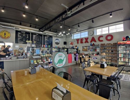 Interior of a casual café with wooden tables, a counter displaying snacks, and a Texaco sign on the wall.