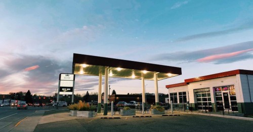 A modern gas station with a bright canopy, surrounded by plants, under a colorful sunset sky.
