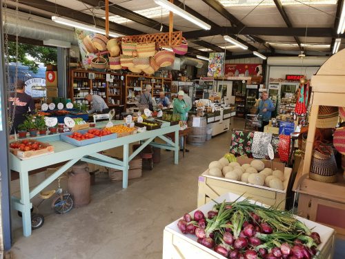 A vibrant market scene with colorful fruits and vegetables displayed on tables, shoppers browsing, and decorative baskets hanging.