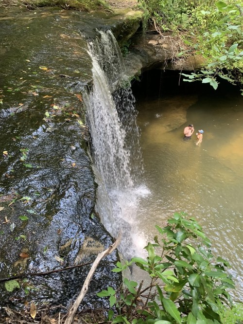 A small waterfall cascades into a serene pool, with two people enjoying the water nearby, surrounded by lush greenery.