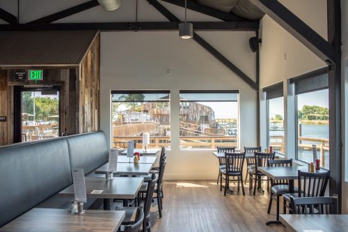 Bright restaurant interior with wooden tables, black chairs, and large windows overlooking a waterfront view.
