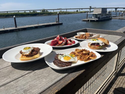 A variety of dishes on a wooden table by the water, including waffles, burgers, and fruit, with a dock in the background.