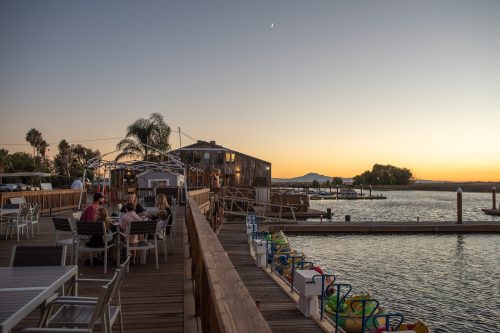 A sunset view of a waterfront restaurant with diners, boats, and colorful paddle boats along the dock.