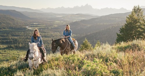 Two women on horseback, overlooking a scenic mountain landscape with lush greenery and distant peaks.