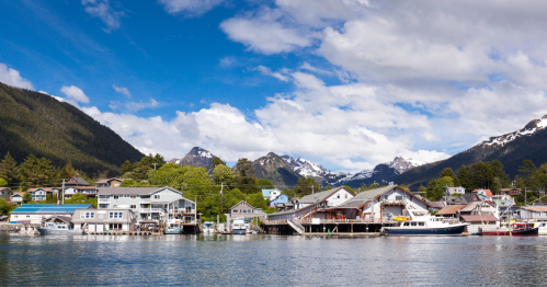 A scenic waterfront view of colorful buildings, boats, and snow-capped mountains under a blue sky with fluffy clouds.