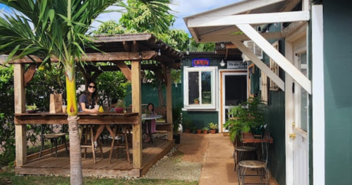 A cozy outdoor café with wooden seating, plants, and an "Open" sign, surrounded by greenery.