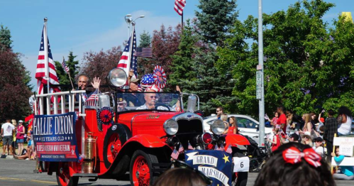 A vintage red car in a parade, decorated with American flags, with two people waving to the crowd.