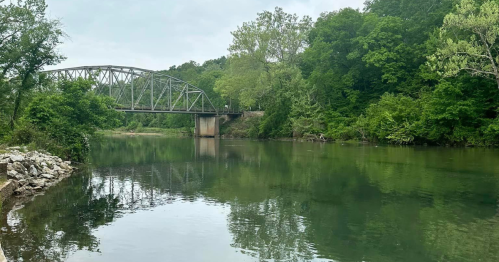 A tranquil river scene with a metal bridge and lush green trees reflecting in the water.