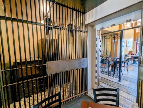 Interior of a restaurant featuring a vintage jail cell with bars, a wooden table, and empty dining area in the background.