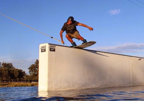 A person performs a wakeboard trick over a water obstacle against a clear blue sky.