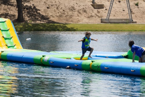 A child in a life jacket jumps on a colorful inflatable water slide, while another person assists nearby in a lake.