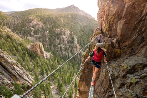 A climber navigates a suspended bridge high above a forested canyon, with mountains in the background.
