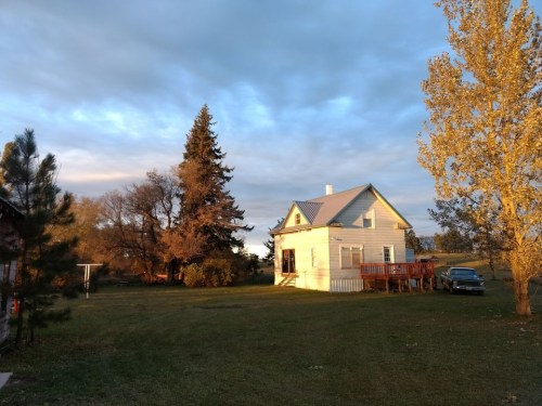A quaint white house with a deck, surrounded by trees and a grassy yard under a cloudy sky at sunset.