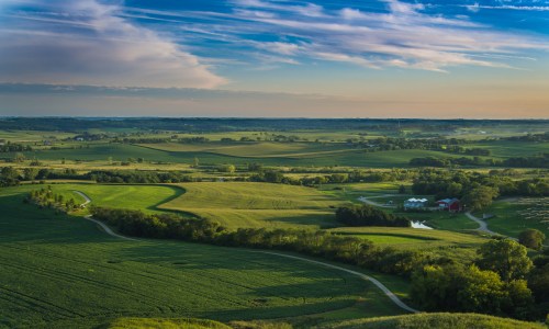 A scenic view of the Wisconsin countryside with rolling green hills, farmland, and scattered trees under a partly cloudy sky.