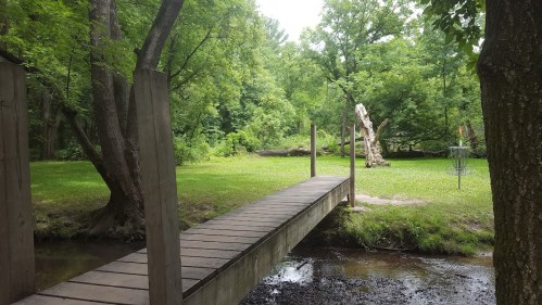 A wooden bridge spans a small stream, surrounded by lush green trees and grass in a serene outdoor setting.