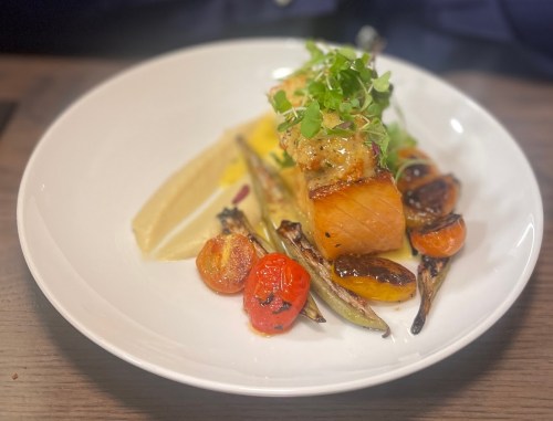 A plated dish featuring salmon, roasted tomatoes, grilled vegetables, and a creamy sauce, garnished with microgreens.