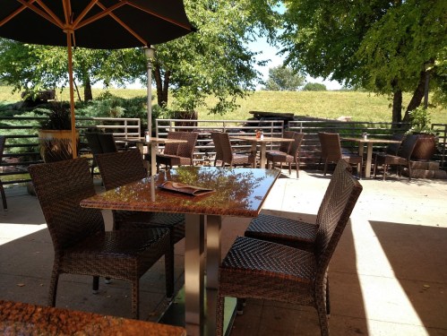 Outdoor dining area with wicker chairs and tables under an umbrella, surrounded by greenery and a sunny sky.