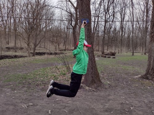 A person in a green jacket and red cap is zip-lining between two trees in a wooded area.