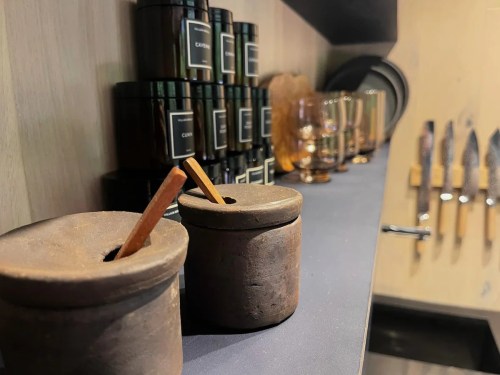 Two small clay containers with wooden sticks, surrounded by candles and kitchen knives on a shelf.