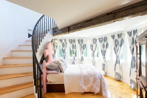 Cozy bedroom with a spiral staircase, large windows, and patterned curtains, featuring a pink headboard and wooden floors.