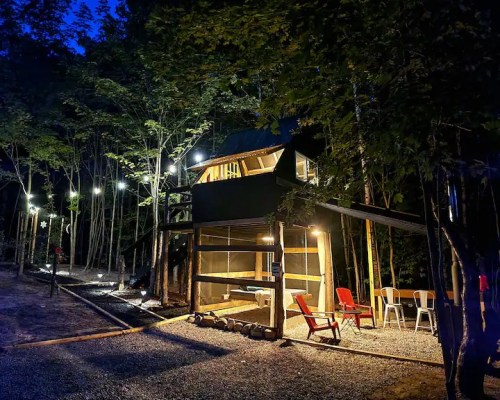 A cozy cabin in the woods at night, illuminated by string lights, with outdoor seating and a gravel path.
