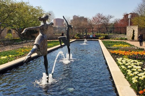 Two dancing sculptures in a garden with a water feature, surrounded by colorful flowers and greenery.